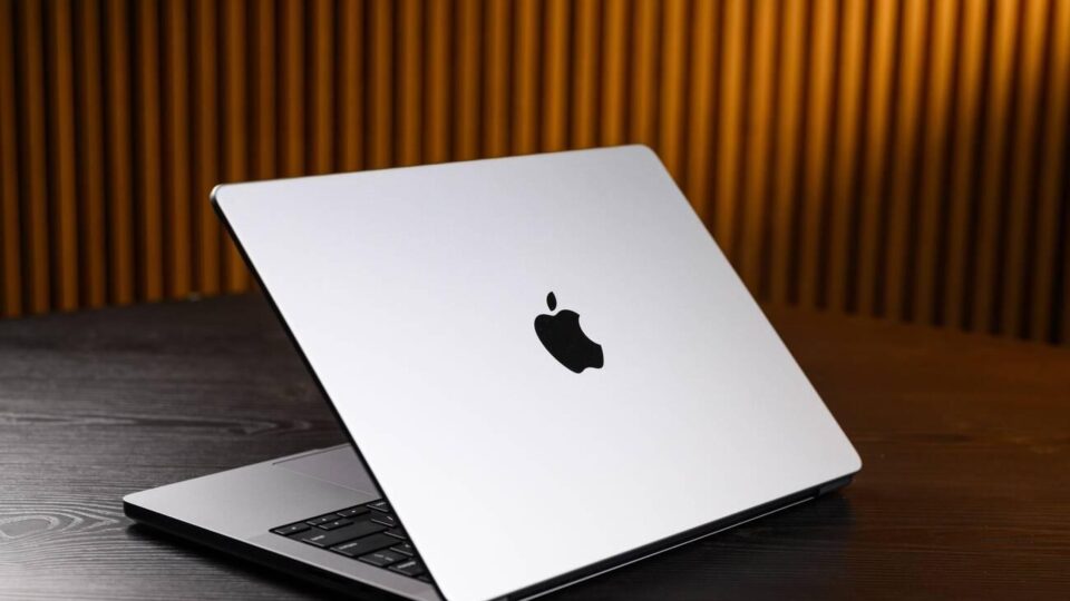 A partially open silver M2 MacBook Pro laptop with a black Apple logo on the back is placed on a dark wooden surface. The background features vertical wooden slats with warm lighting. This image shows a sleek, partially open silver Apple laptop on a dark wooden surface. The contrast between the laptop's metallic finish and the dark wood, along with the warm lighting from the vertical wooden slats in the background, creates a visually appealing and professional setting. This image is relevant for showcasing modern technology and design aesthetics.